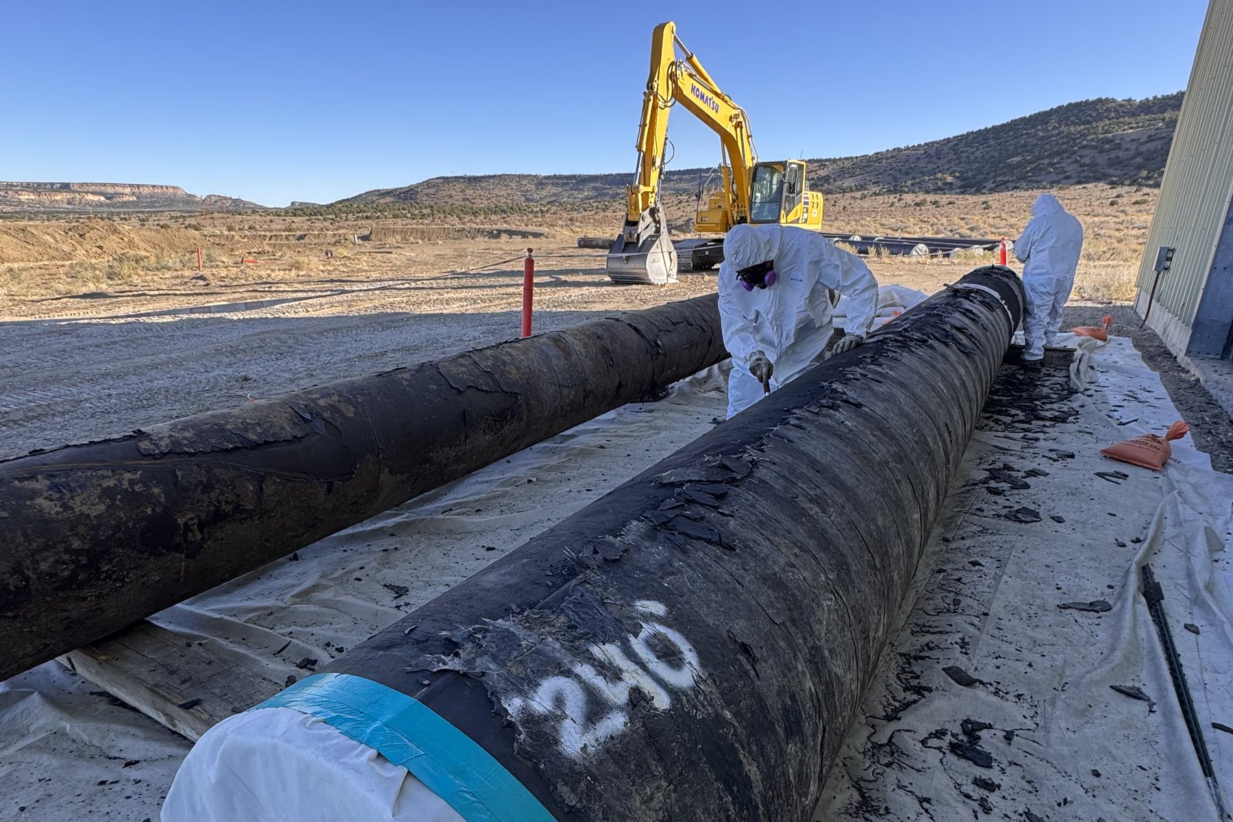 DEKON technicians in Tyvek PPE performing asbestos pipe-wrap abatement with Komatsu excavator support