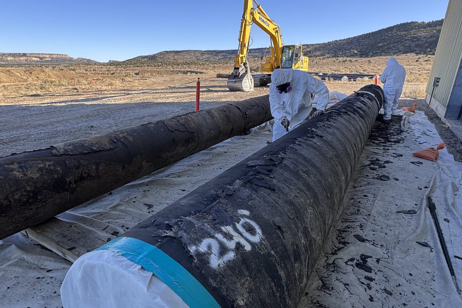 DEKON crew in Tyvek performing wet-method asbestos pipe wrap abatement on an excavated underground steel pipe